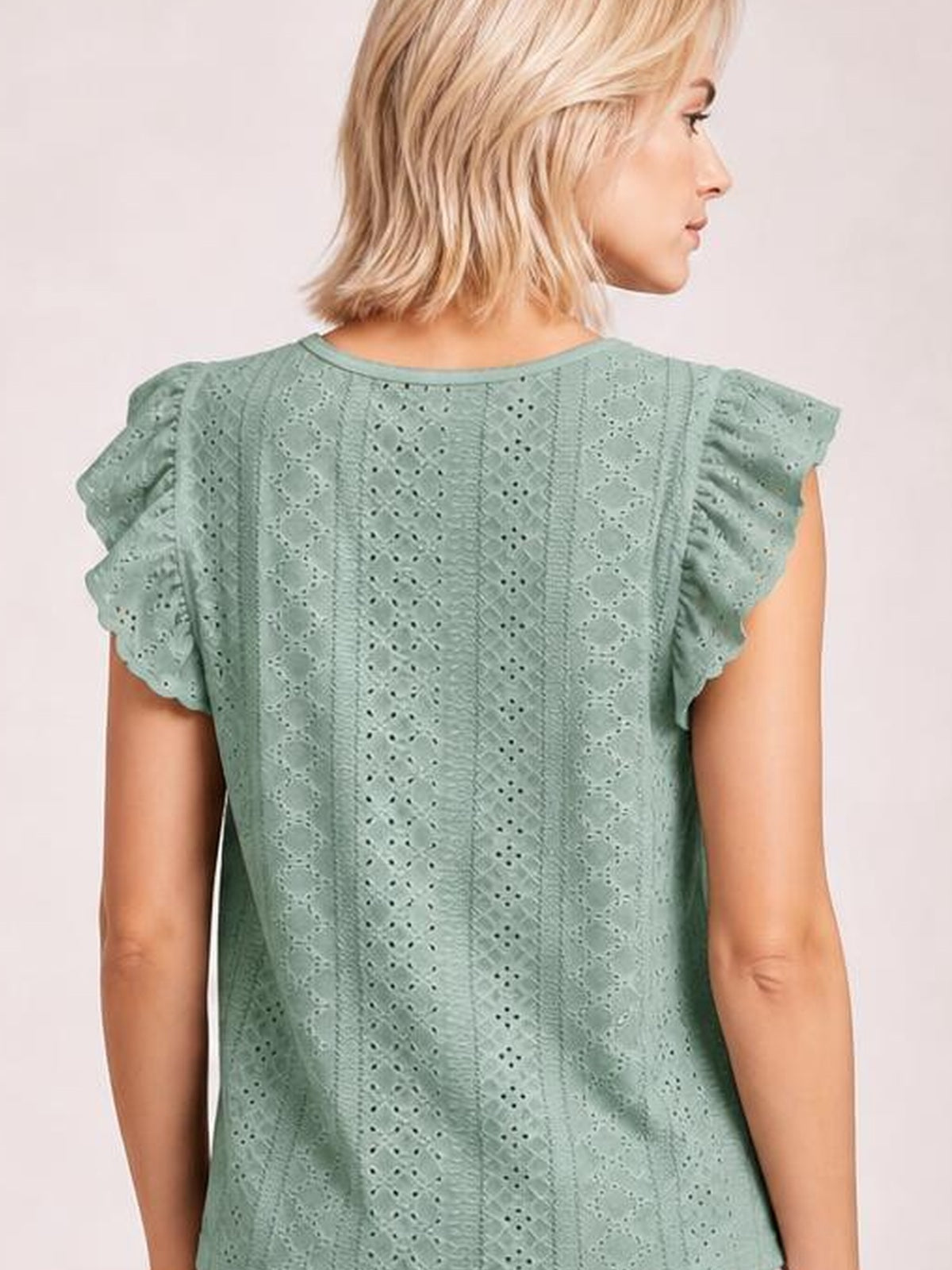 A model shown from behind wears a sage green eyelet top with a round neck, ruffled cap sleeves, vertical cutout panels, and a relaxed straight silhouette.