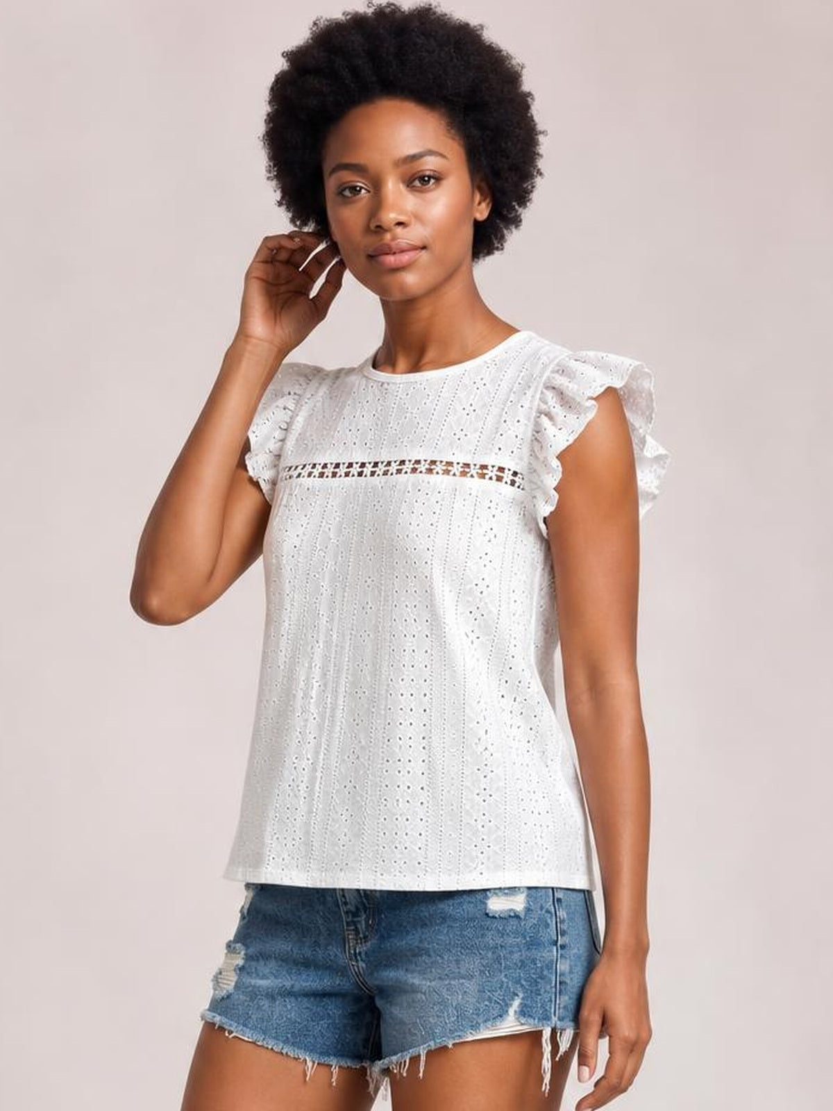 Model wearing a white eyelet round neck cap sleeve top with ruffled sleeves, a horizontal cutout band, and a relaxed straight silhouette, posing with one hand near her ear.