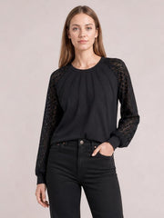 A model stands in a black eyelet round neck long sleeve blouse with semi-sheer patterned sleeves, gathered neckline pleats, and a relaxed silhouette.