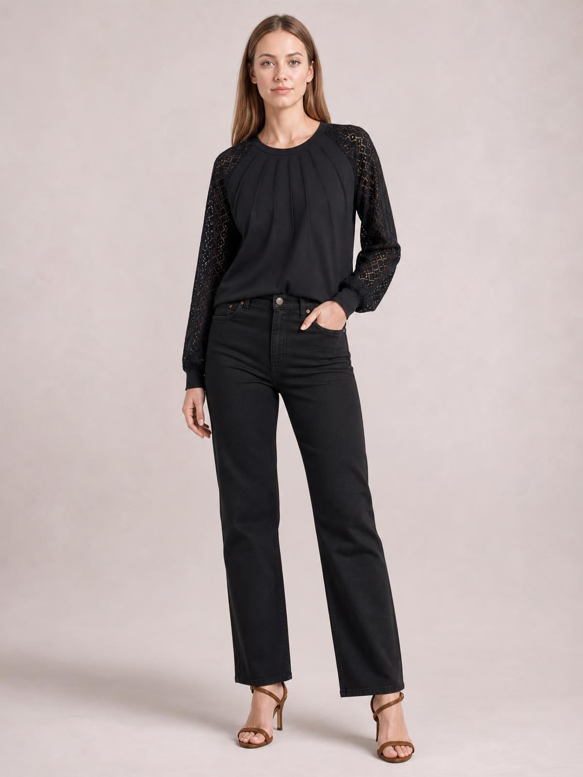 A model wears a black round-neck long-sleeve blouse with eyelet semi-sheer sleeves, pleated front detailing, and a relaxed silhouette, posing with one hand in her pocket.