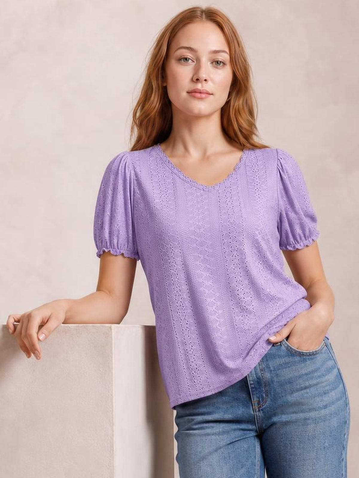 A model wears a lavender eyelet blouse with short gathered sleeves and delicate frill edging, posing with one hand on a ledge and the other in her jeans pocket.