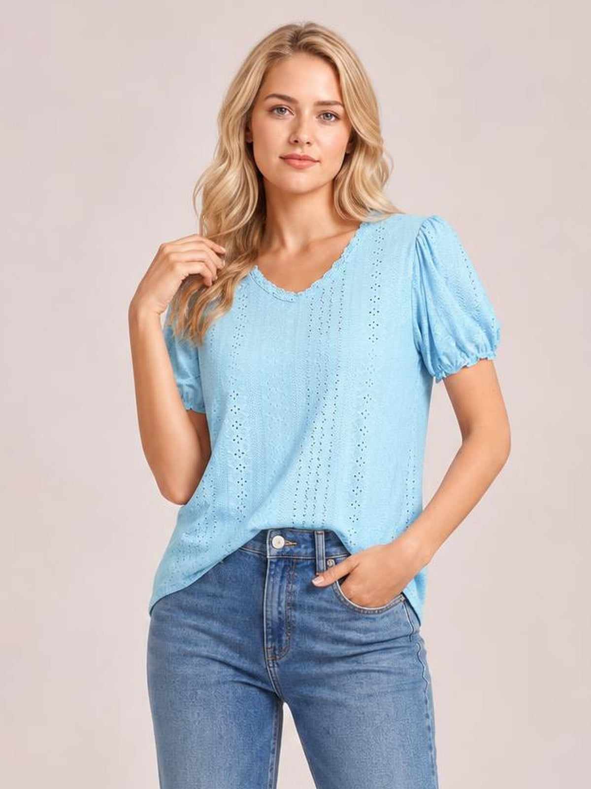 Model wearing a light blue eyelet blouse with a round frilled neckline, puffed short sleeves, and a relaxed silhouette, posing with one hand in her jeans pocket.