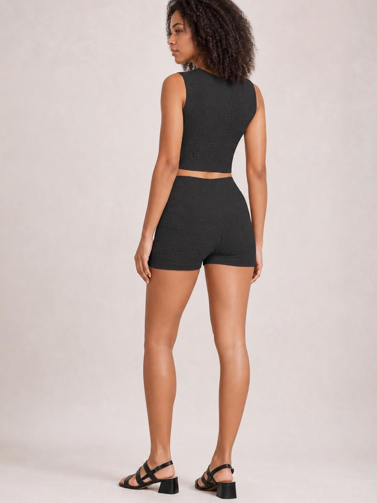 Model stands in a back view wearing a black eyelet round neck tank and matching high-waisted sweater shorts with ribbed hems and a fitted silhouette.