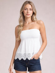 A model wears a white eyelet smocked tube top with a strapless neckline, flowing silhouette, and scalloped cutout hem, paired with denim shorts.