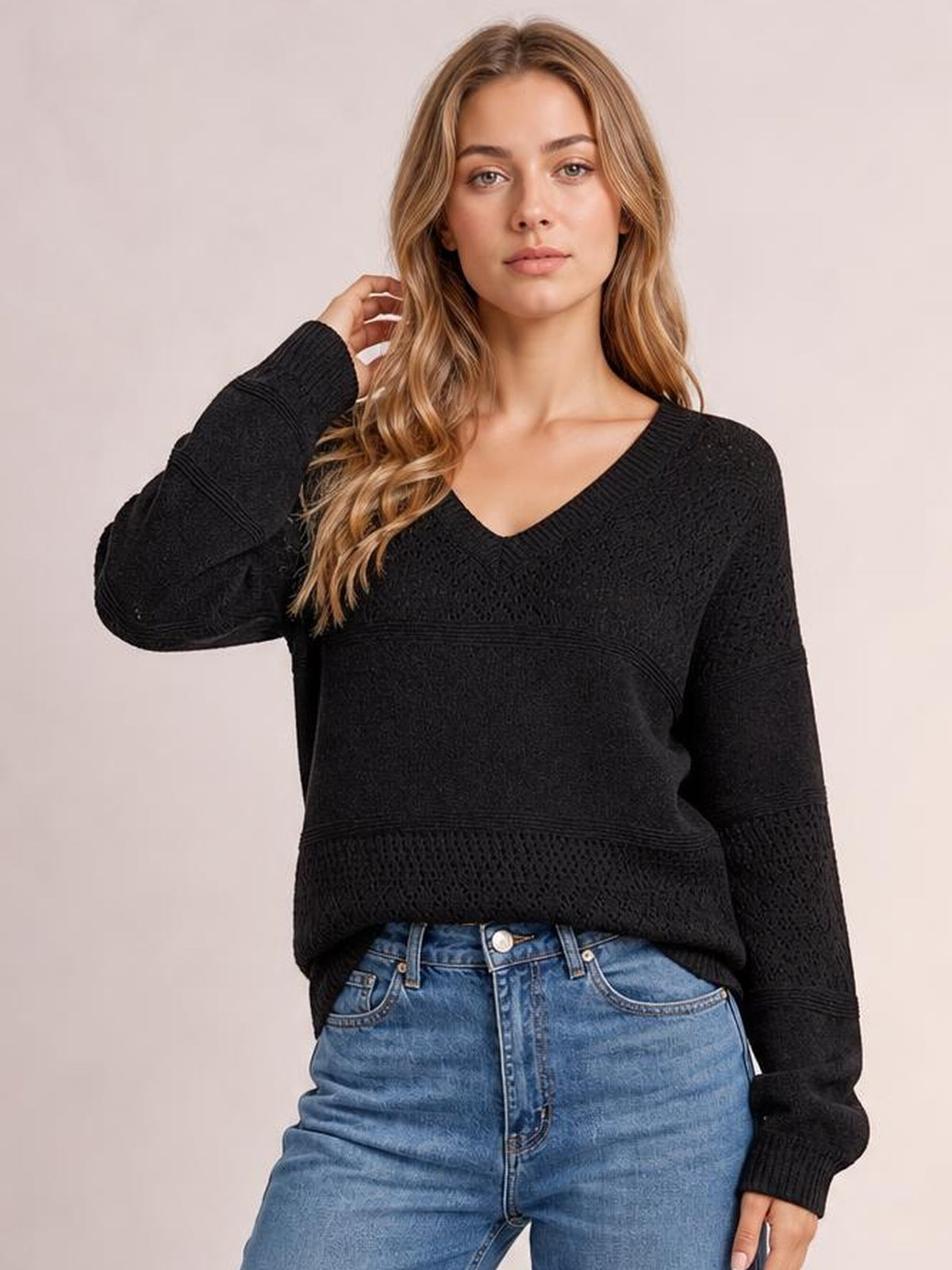 A model poses with one hand near her hair, wearing a black eyelet V-neck long-sleeve sweater with a relaxed silhouette and ribbed cuffs.