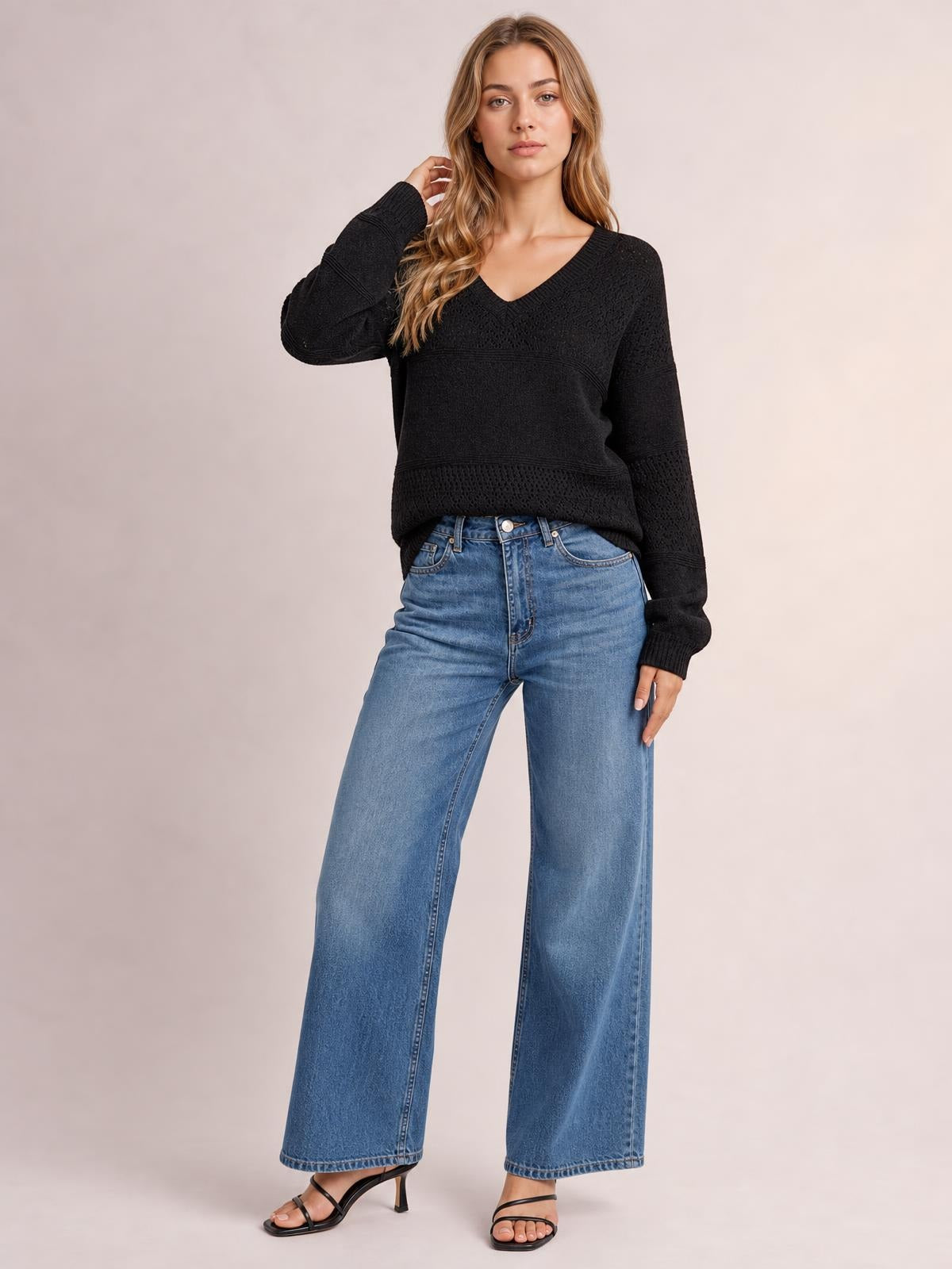 Model poses in a black eyelet V-neck long sleeve sweater with a relaxed silhouette, ribbed cuffs, and textured panel construction.