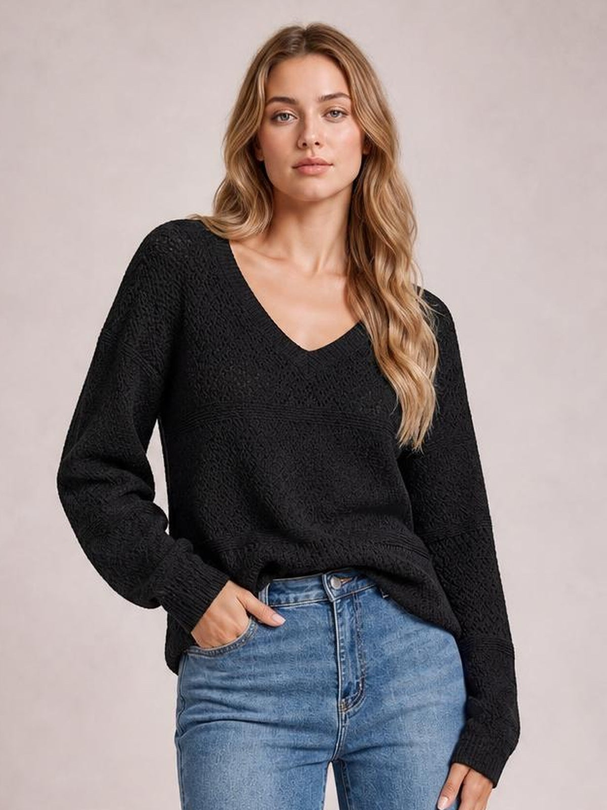 Model wearing a black eyelet V-neck long sleeve sweater with a relaxed silhouette and ribbed cuffs, one hand tucked into her jeans pocket.