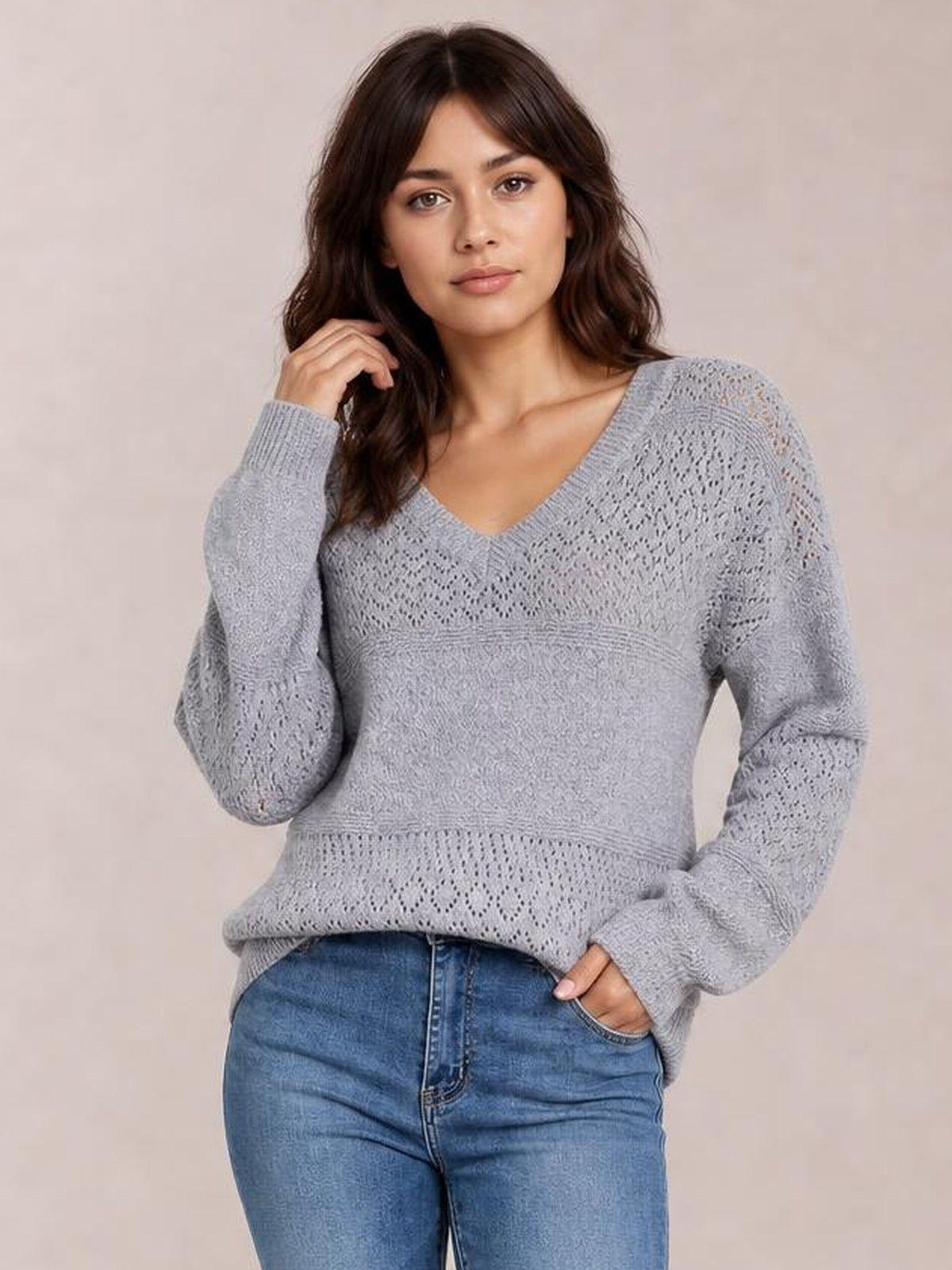 Model poses in a light gray eyelet V-neck long sleeve sweater with an openwork knit pattern, ribbed cuffs and hem, and a relaxed silhouette.
