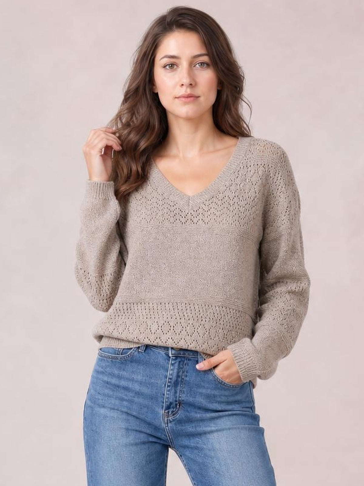 Model wearing a taupe eyelet V-neck long sleeve sweater with a relaxed silhouette, ribbed cuffs and hem, posing with one hand in her jeans pocket.