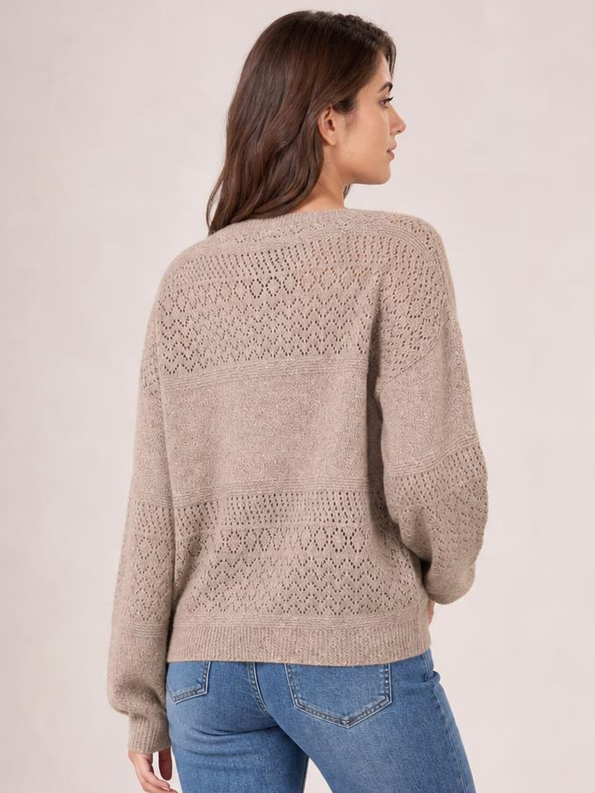 A model turned slightly away wears a taupe eyelet long-sleeve sweater with an open-knit back, relaxed silhouette, and ribbed hem and cuffs.