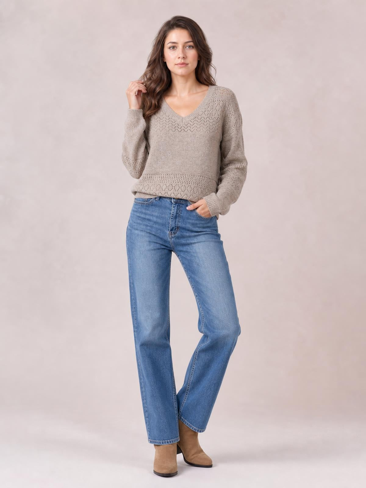 Model wears a taupe eyelet V-neck long sleeve sweater with a relaxed silhouette and ribbed trim, posing with one hand in her jeans pocket.
