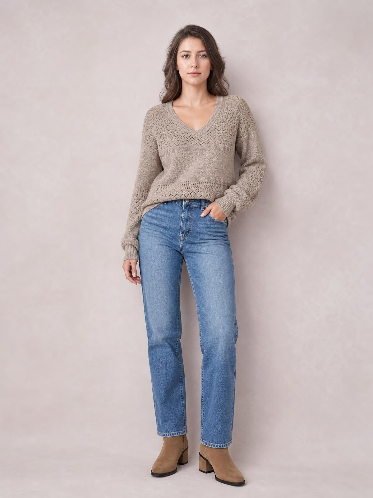 Model poses in a taupe eyelet V-neck long sleeve sweater with a relaxed silhouette, ribbed cuffs and hem, paired with jeans.