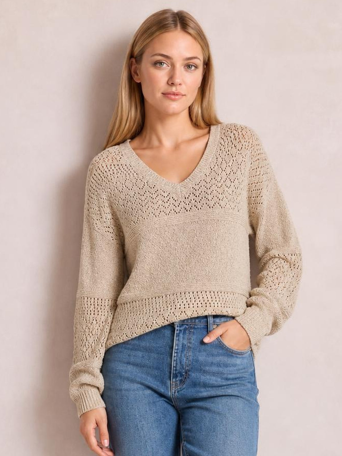 A woman models a beige eyelet V-neck long sleeve sweater with an openwork knit pattern, relaxed silhouette, ribbed cuffs, and one hand in her jeans pocket.