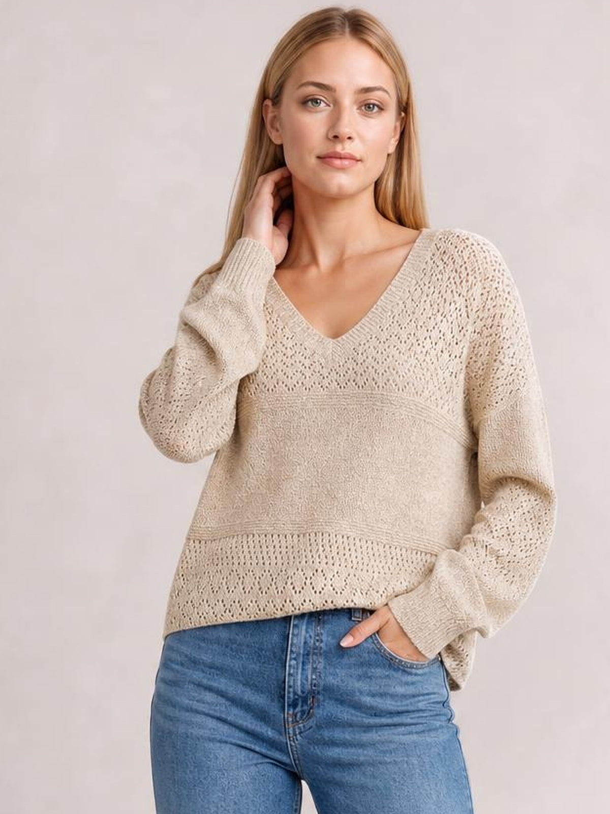 A model poses in a cream eyelet V-neck long sleeve sweater with openwork knit panels, ribbed trim, and a relaxed silhouette, one hand at her neck.