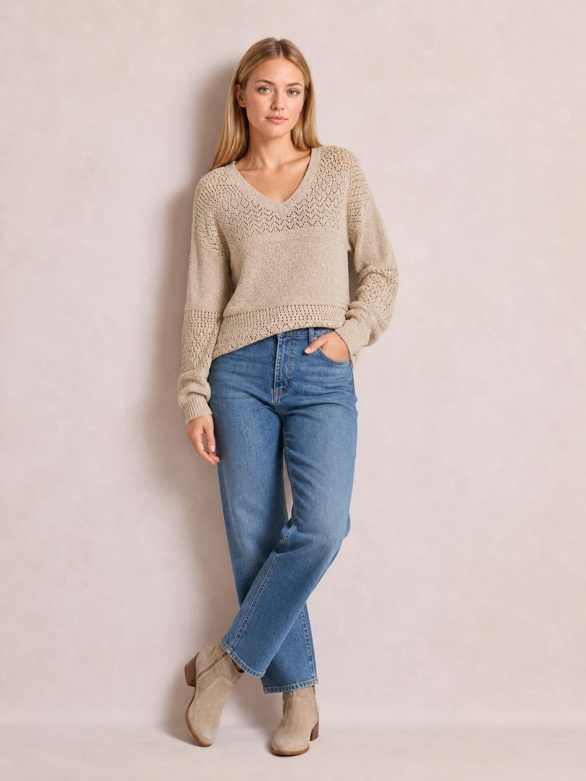 Model wears a beige eyelet V-neck long sleeve sweater with a relaxed silhouette, open-knit panels, ribbed cuffs and hem, and one hand in her jeans pocket.