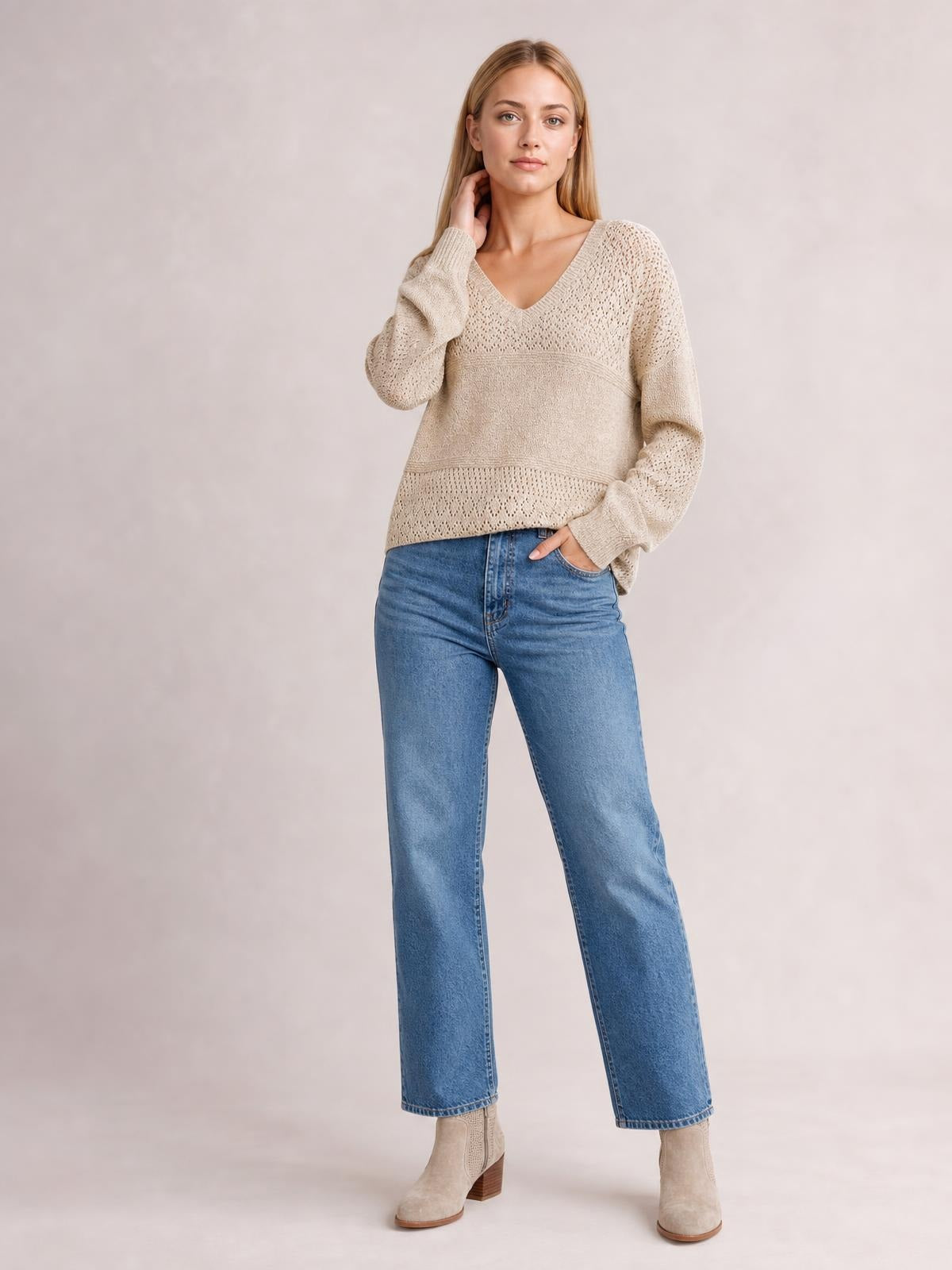 Model wears a beige eyelet V-neck long sleeve sweater with a relaxed silhouette, textured knit panels, and ribbed cuffs and hem, posing with one hand in her pocket.