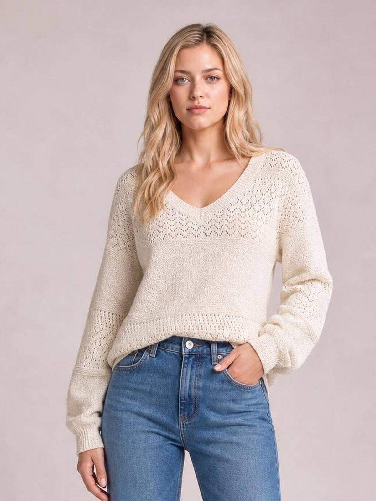 Model wears an ivory eyelet V-neck long sleeve sweater with a relaxed silhouette, ribbed cuffs and hem, shown with one hand in her jeans pocket.
