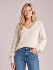 Model wears an ivory eyelet V-neck long sleeve sweater with a relaxed silhouette, ribbed cuffs and hem, shown with one hand in her jeans pocket.