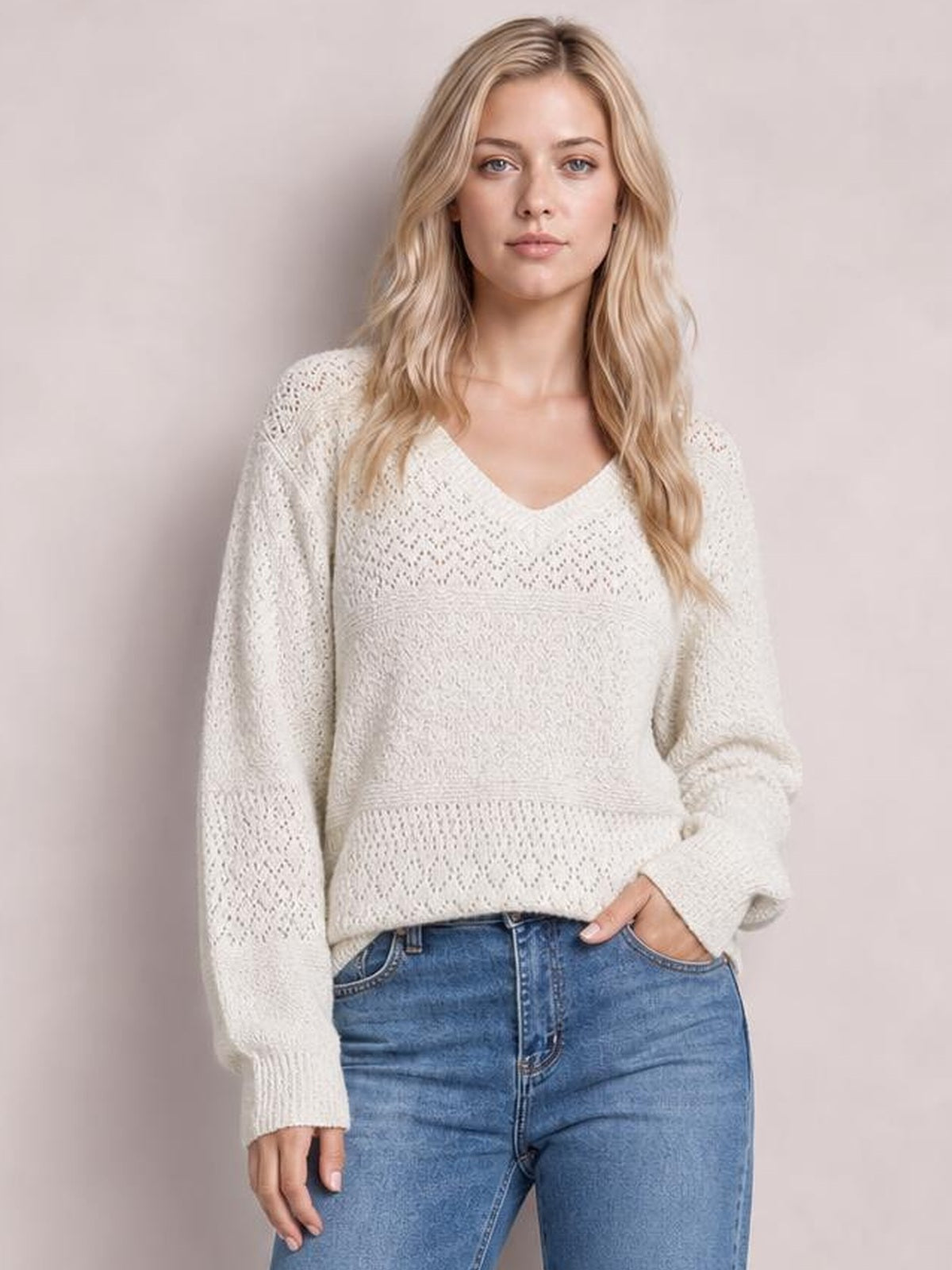 Model wears a cream eyelet V-neck long sleeve sweater with a relaxed silhouette, textured knit panels, ribbed cuffs and hem, and one hand in her jeans pocket.