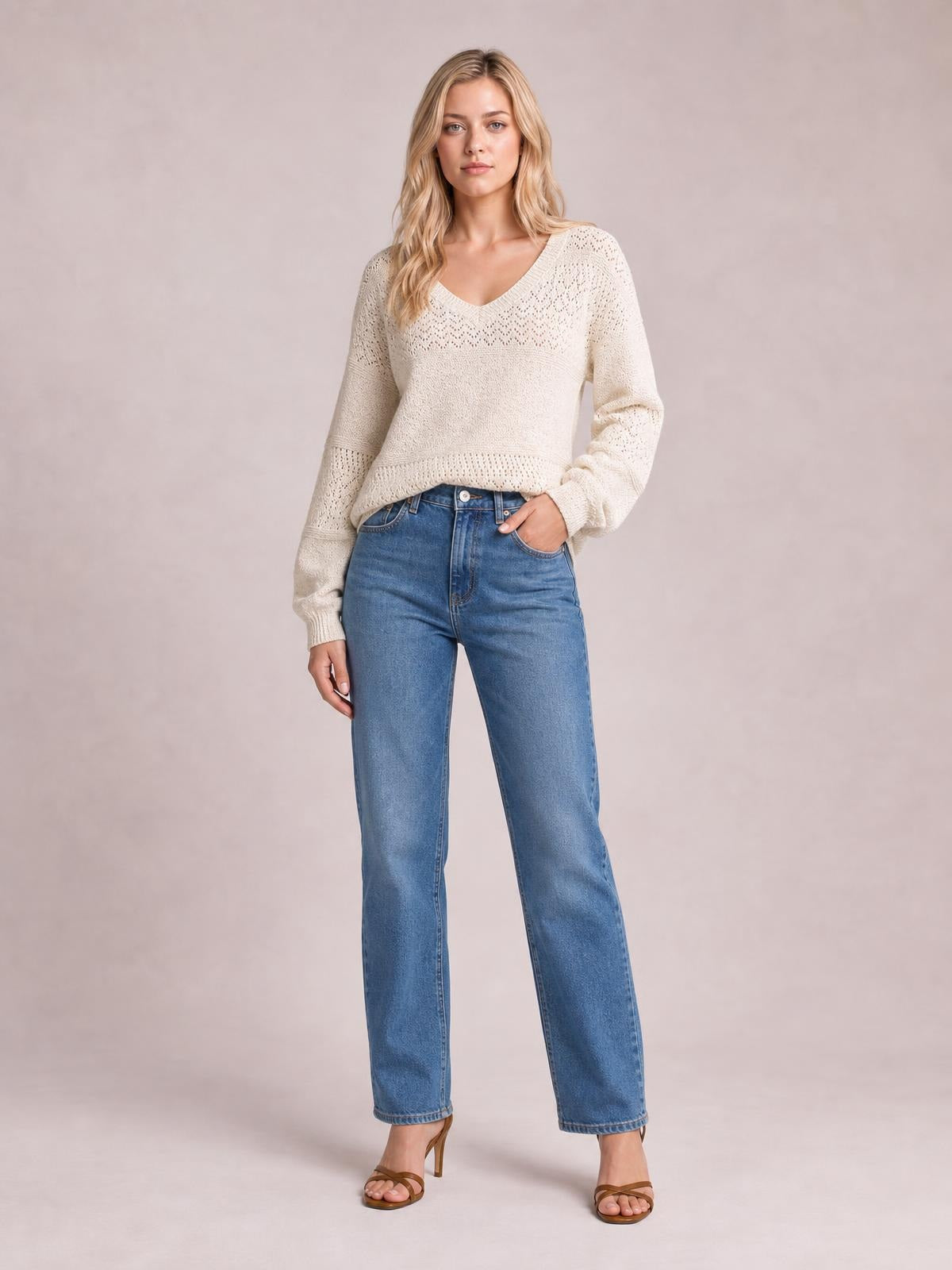A model stands with one hand in her jeans pocket, wearing a cream eyelet V-neck long sleeve sweater with a relaxed silhouette, ribbed cuffs, and hem.