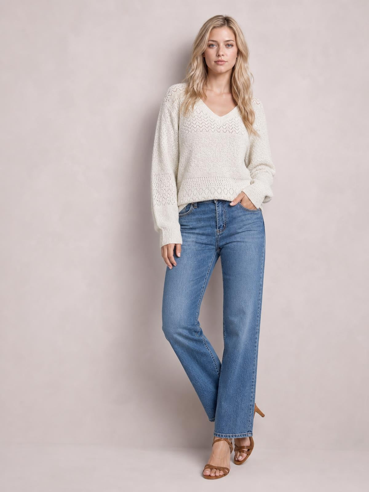 A model wears an ivory eyelet V-neck long sleeve sweater with a relaxed silhouette, textured open-knit panels, ribbed cuffs and hem, and blue jeans.