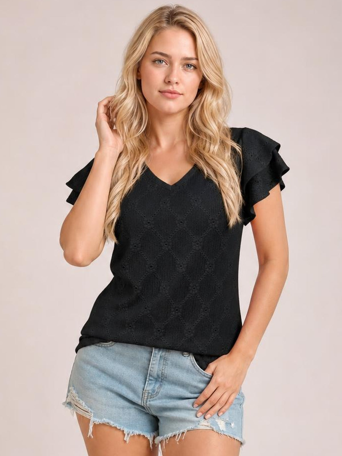 Model wears a black eyelet lace top with a V-neck, ruffled cap sleeves, and a relaxed silhouette, posing with one hand in her pocket.