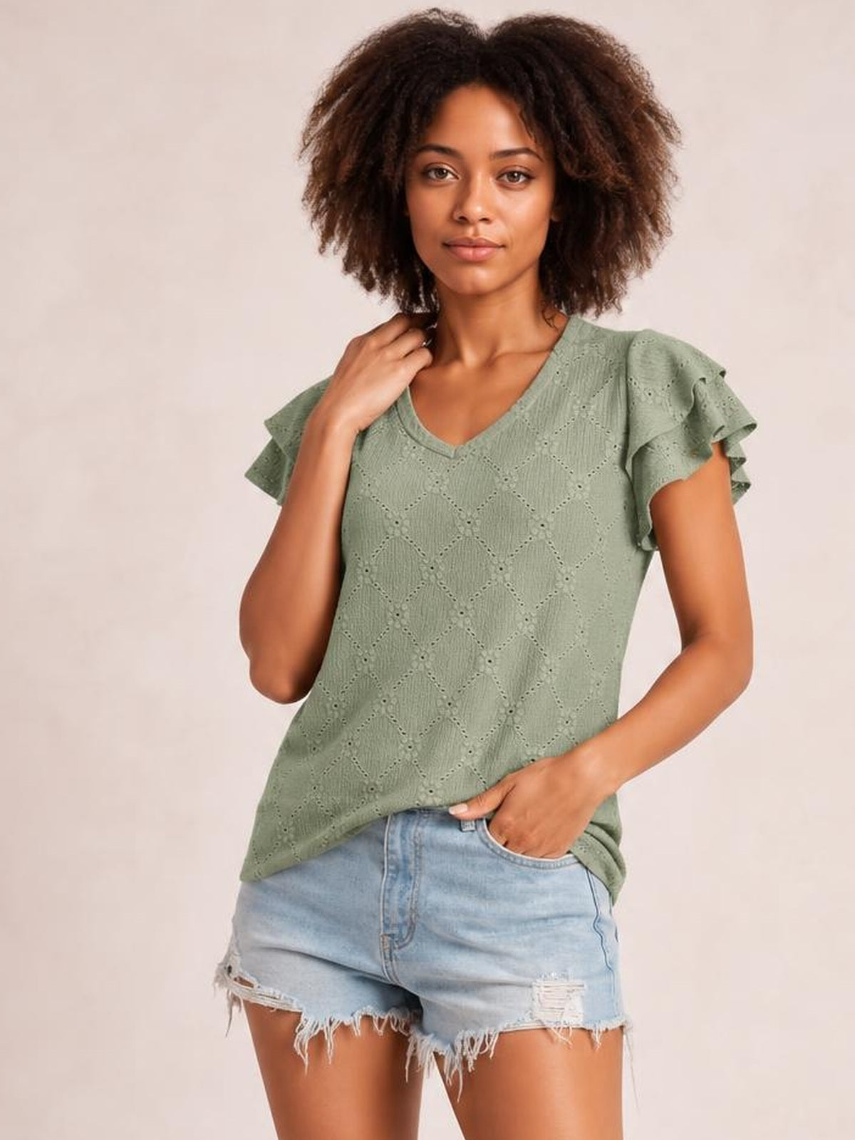 Model poses in a sage green eyelet top with a V-neck, layered ruffled cap sleeves, lace-like cutout texture, and a relaxed silhouette.