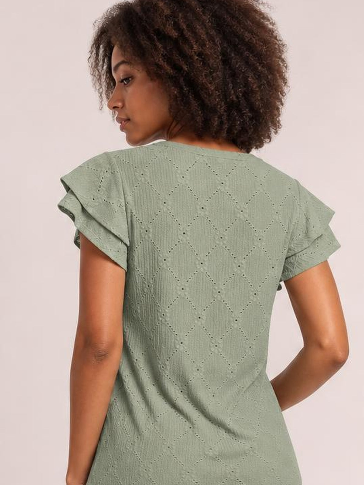 Model shown from behind wearing a sage green eyelet top with a relaxed silhouette, decorative lace pattern, and layered ruffled cap sleeves.