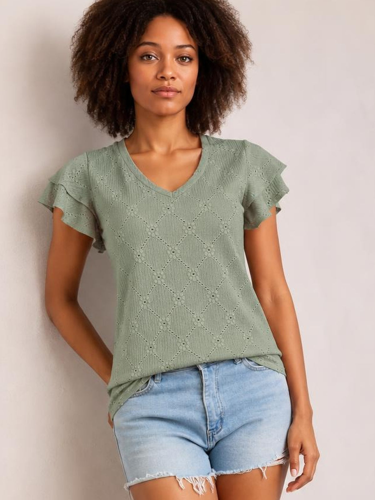 Model poses with one hand in her pocket wearing a sage green eyelet V-neck top with ruffled cap sleeves and a relaxed straight silhouette.