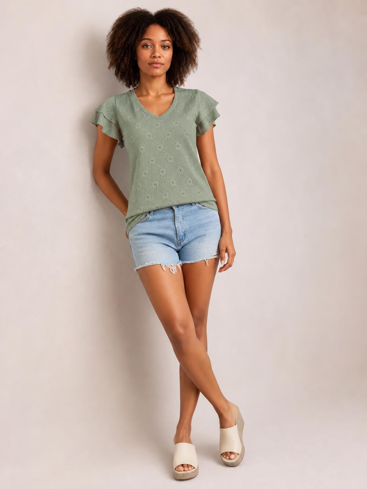 Model poses with one hand in her pocket wearing a sage green eyelet V-neck top with ruffled cap sleeves and a relaxed straight silhouette.