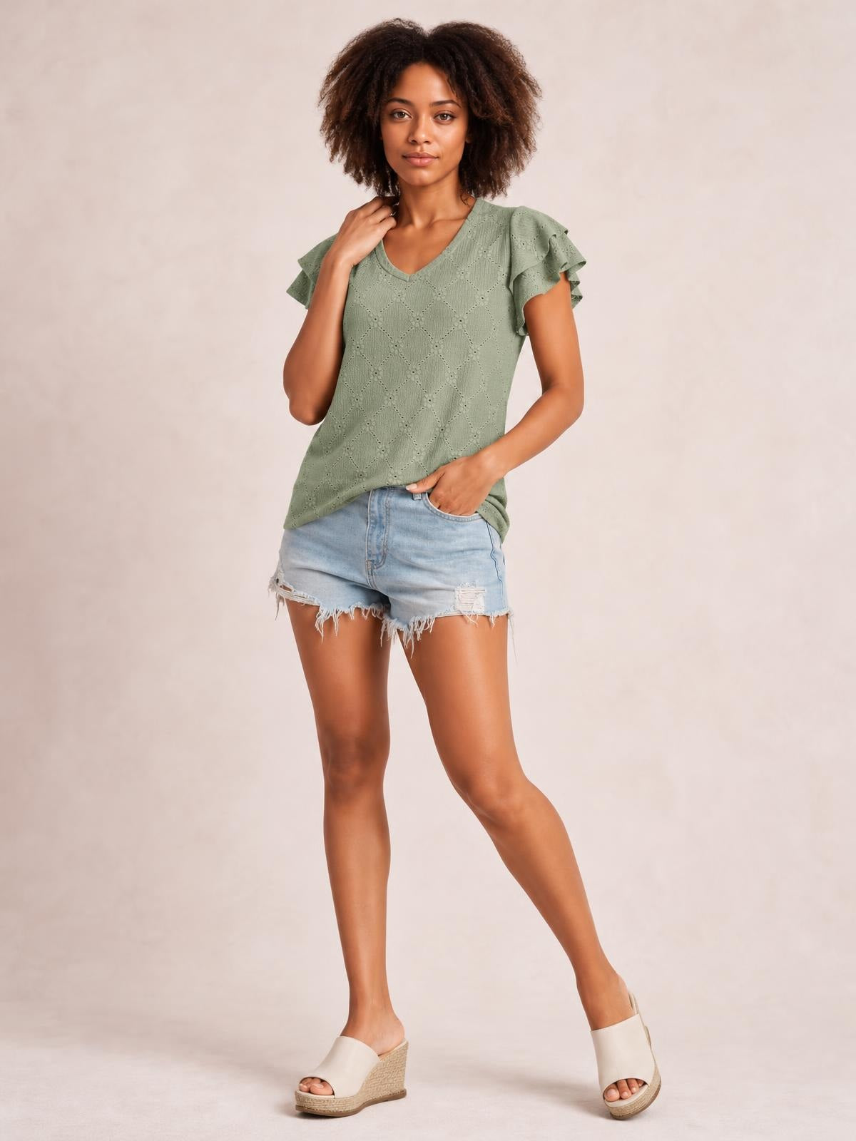 A model poses in a sage green eyelet top with a V-neck, layered ruffled cap sleeves, lace-like cutout texture, and a relaxed straight silhouette.