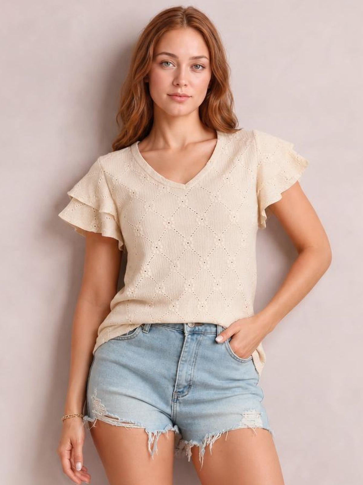 A model poses in a cream eyelet top with a V-neck, delicate lace texture, layered ruffled cap sleeves, and a relaxed straight silhouette.
