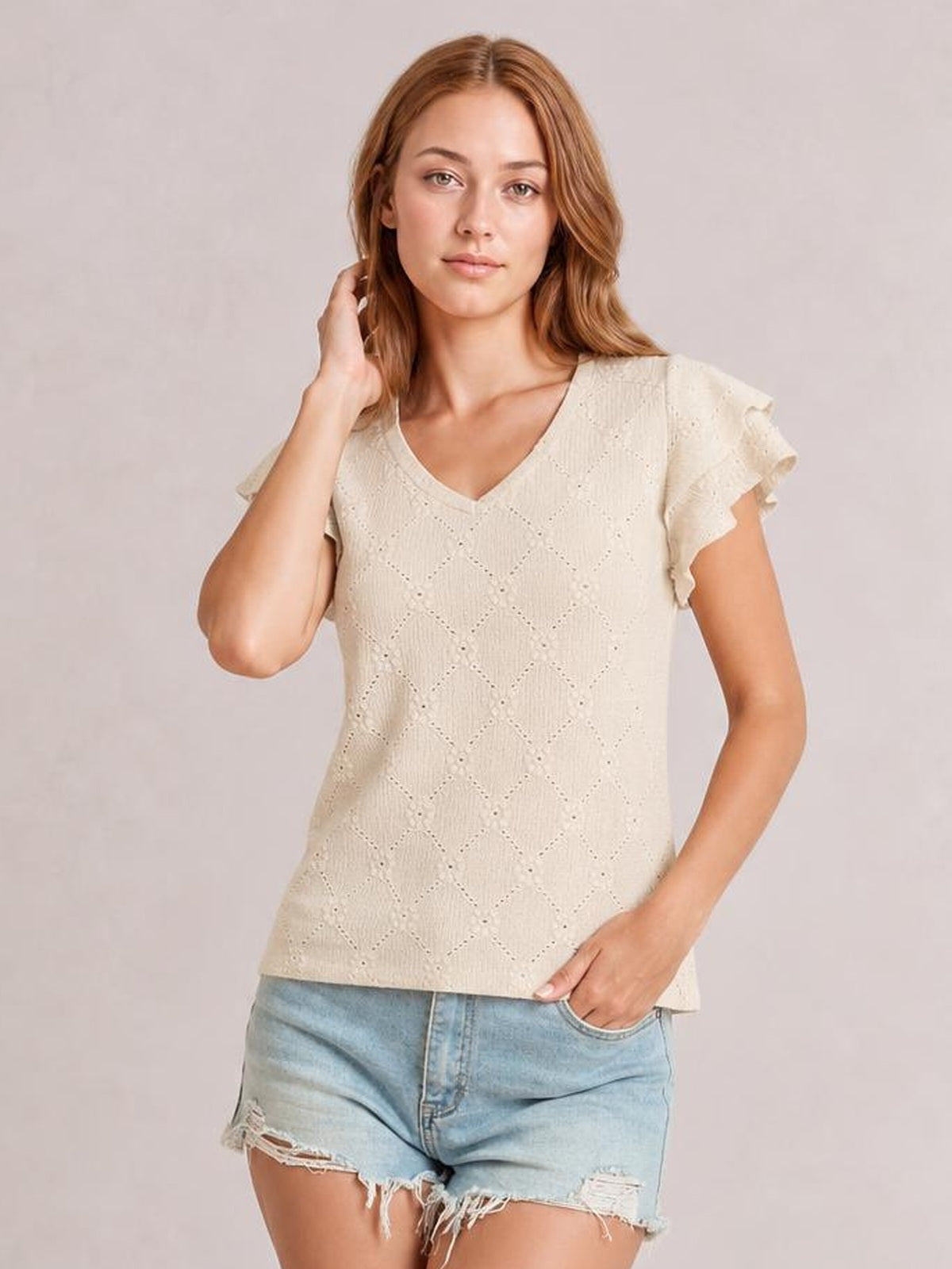 A model poses in a cream eyelet top with a V-neck, ruffled cap sleeves, lace-like detailing, and a relaxed straight silhouette.
