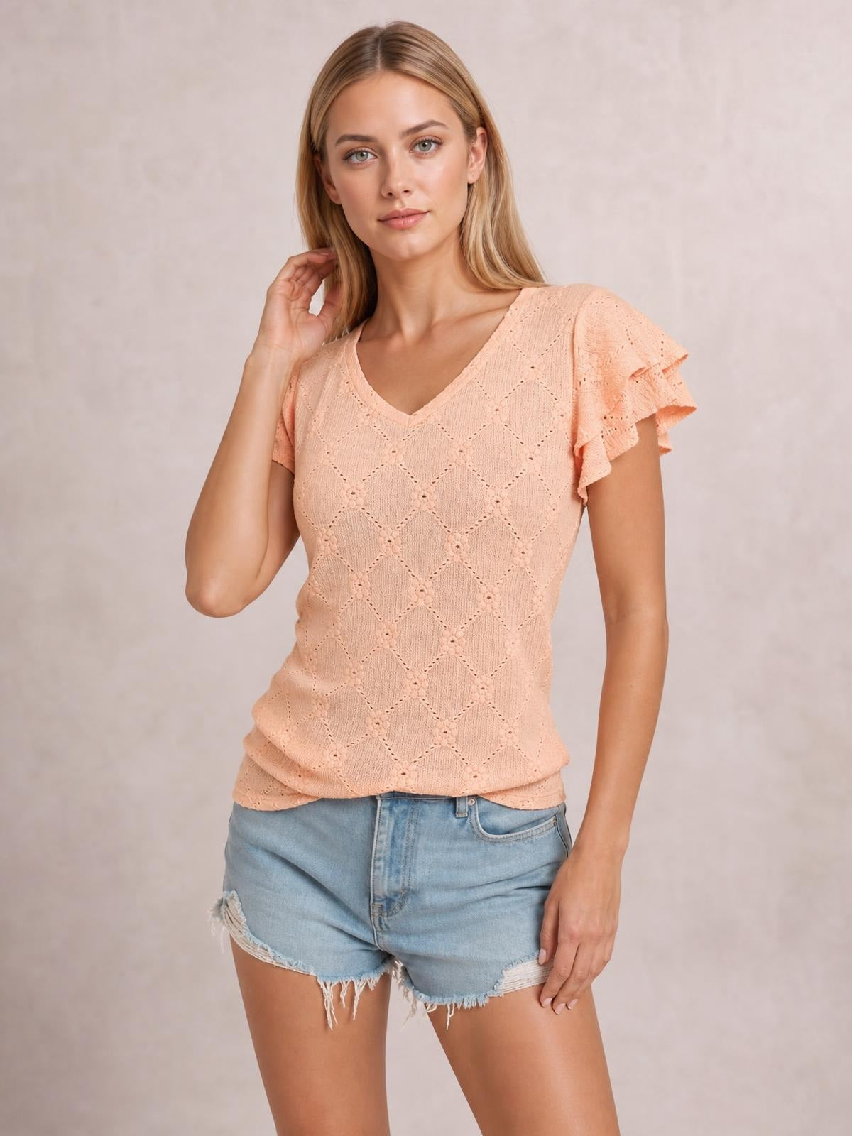 Model poses with one hand in her hair wearing a peach eyelet lace top with a V-neck, ruffled cap sleeves, and a fitted silhouette.