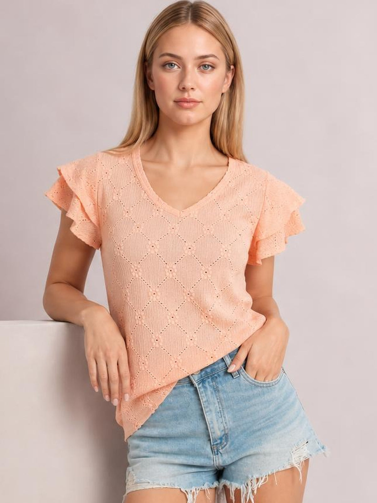 Model wears a peach eyelet top with a V-neck, layered ruffled cap sleeves, lace texture, decorative buttons, and a relaxed silhouette.