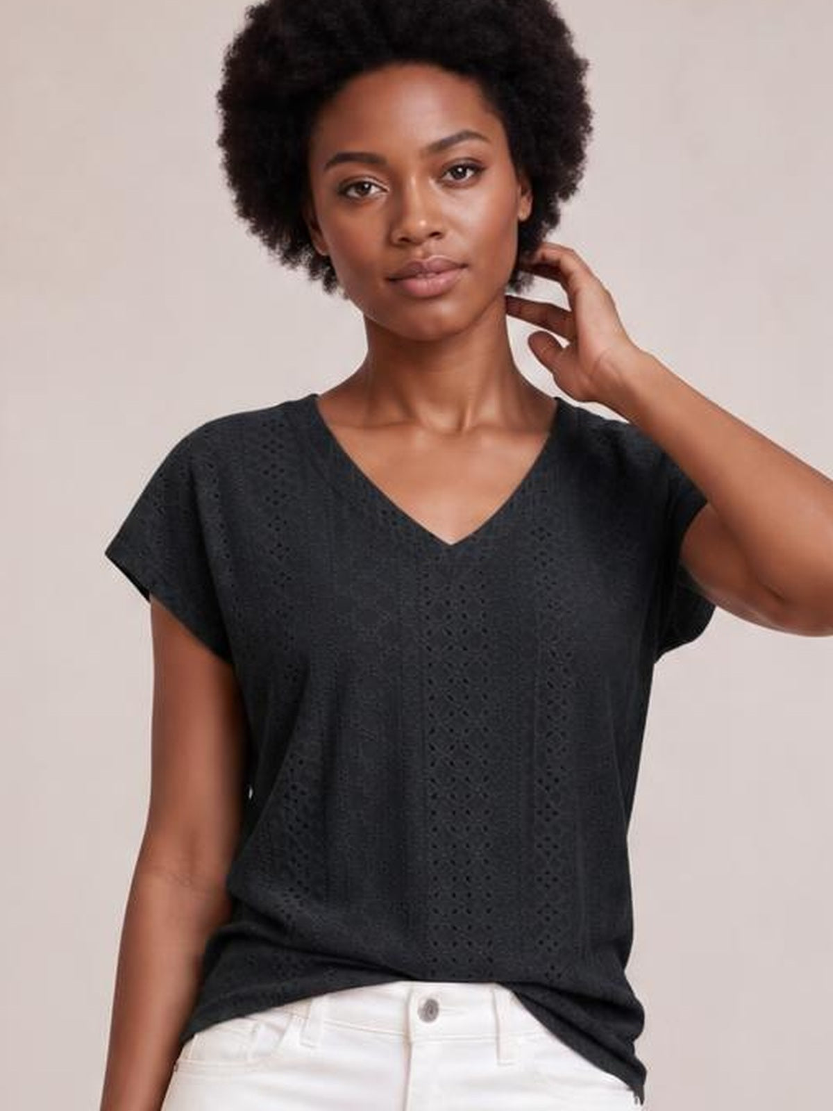 Model wearing a black eyelet V-neck short sleeve top with a relaxed silhouette, shown from the waist up while touching her neck.