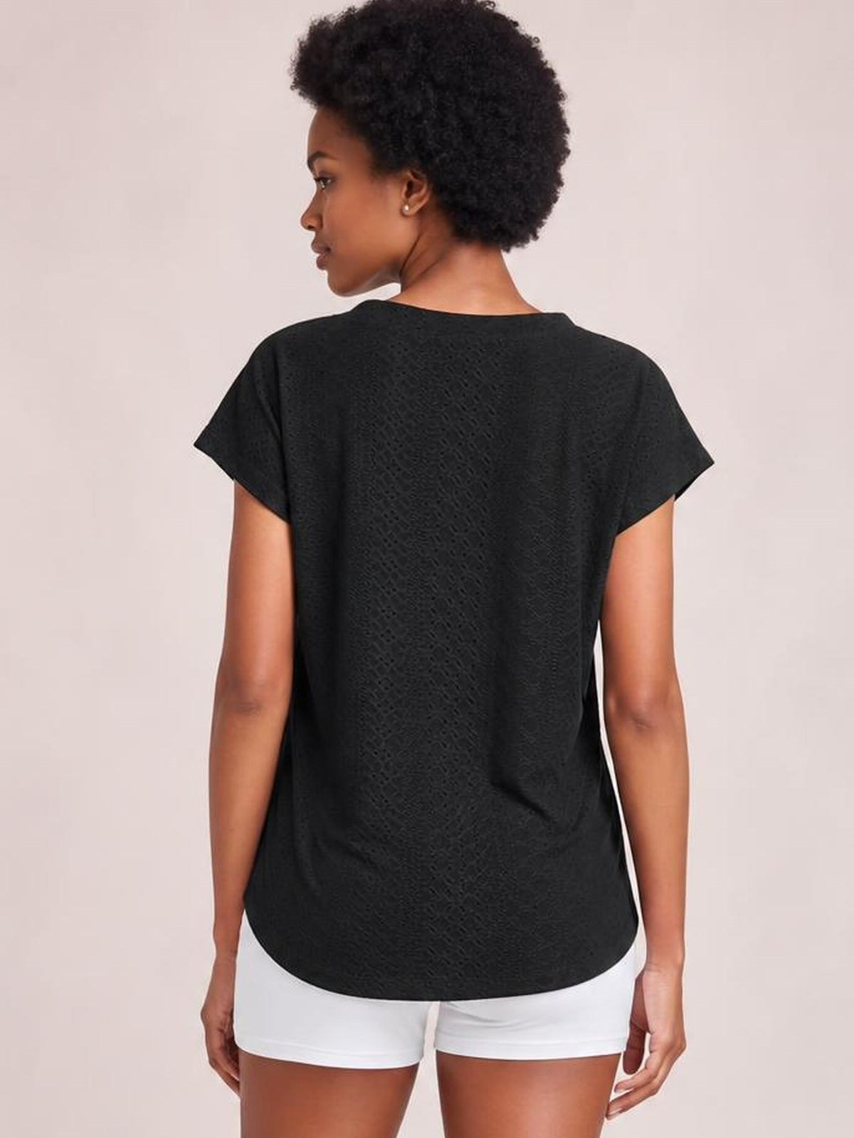 Model shown from behind wearing a black eyelet short-sleeve top with a relaxed silhouette, softly rounded hem, and slightly loose construction.