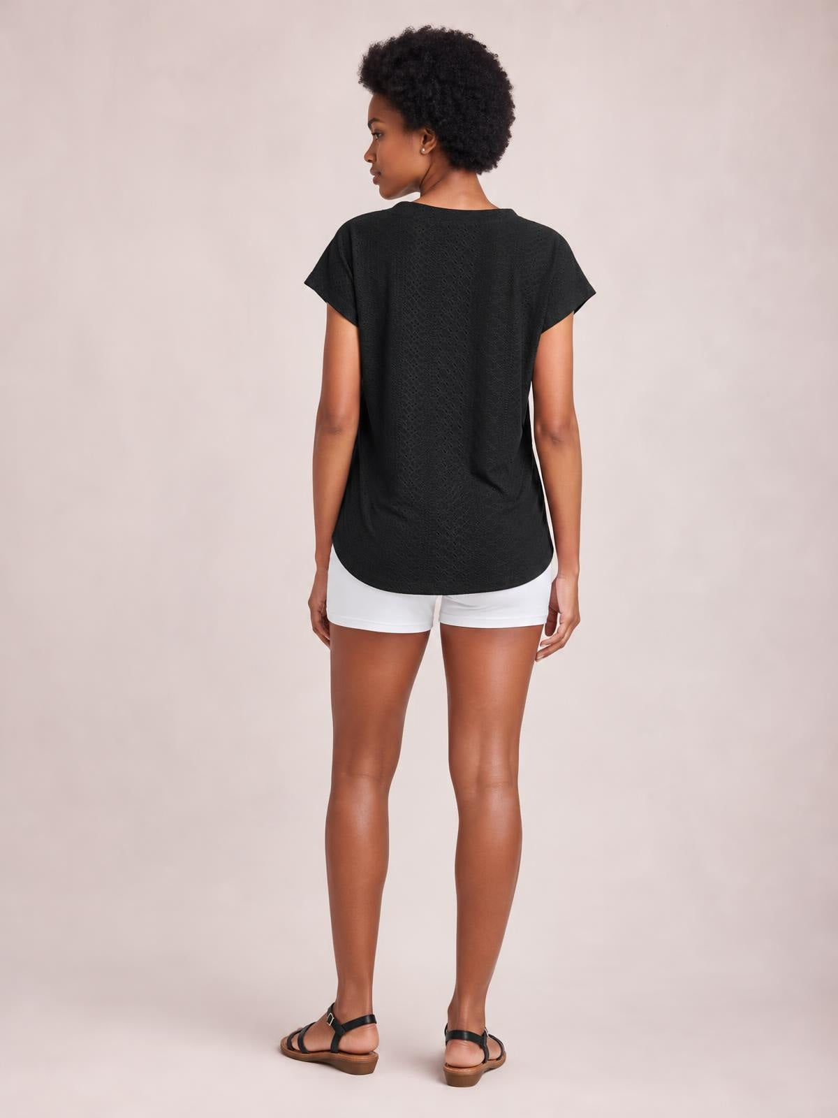 Model shown from behind wears a black eyelet short sleeve top with a relaxed straight silhouette, rounded hem, and subtly textured construction.