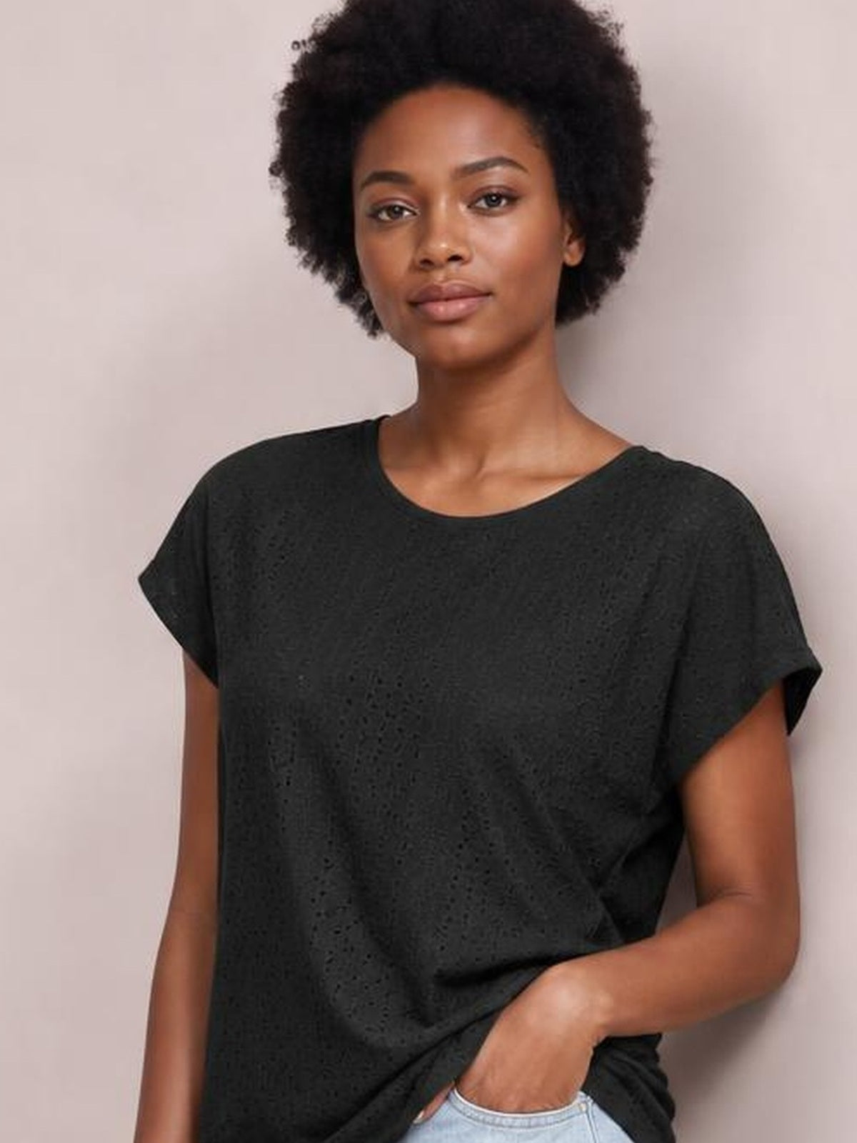 A model wears a black eyelet short-sleeve top with a relaxed silhouette and rounded neckline, posing with one hand tucked into her jeans pocket.
