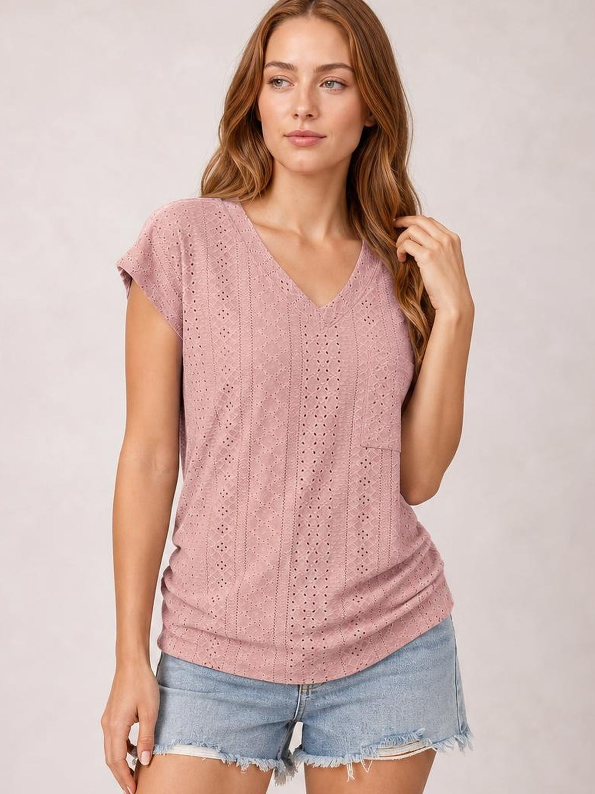 A model poses in a dusty rose eyelet V-neck short-sleeve top with a relaxed silhouette, subtle side pocket, and slightly stretchy construction.