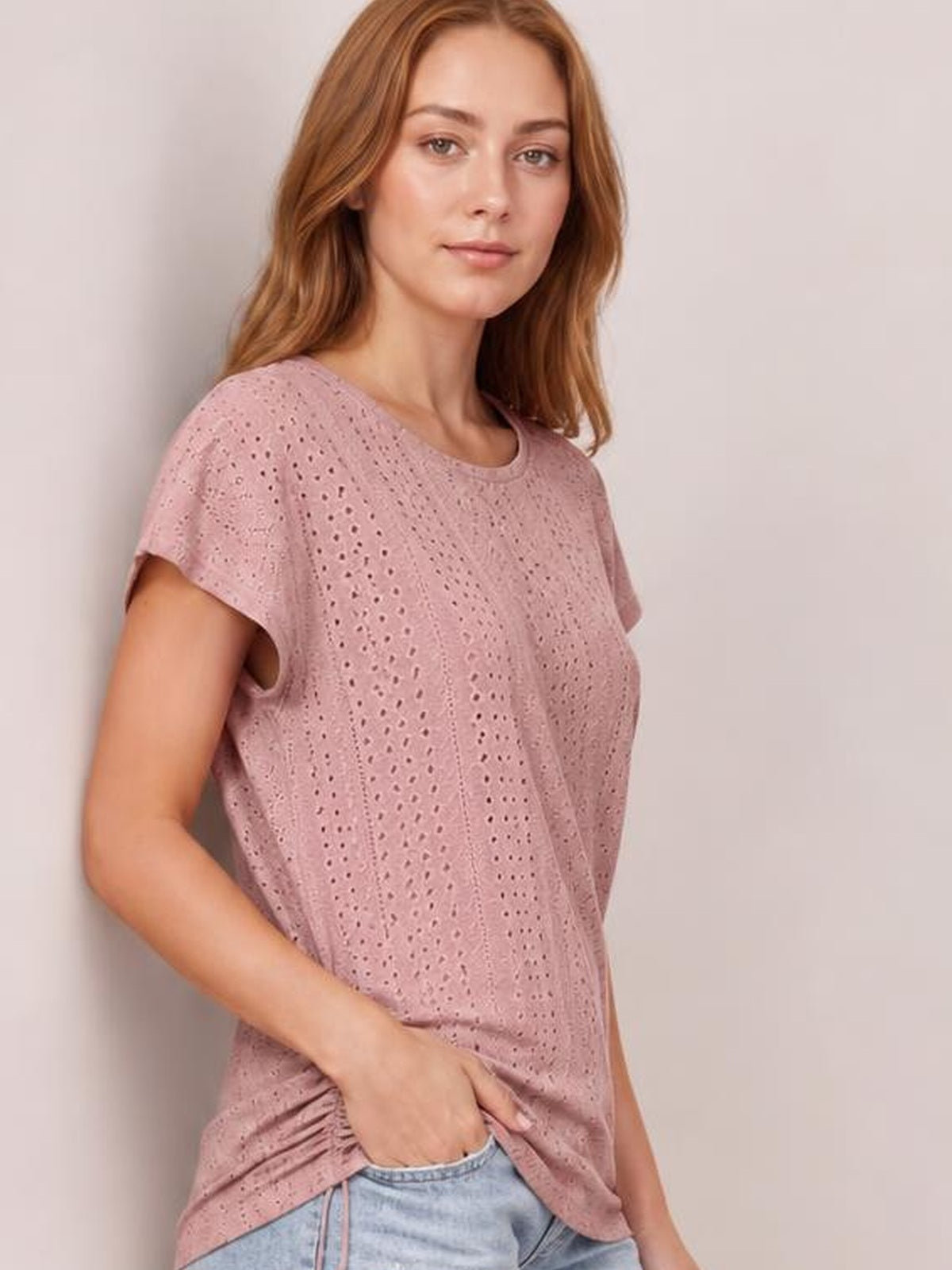 A model wears a dusty pink eyelet short sleeve top with a relaxed silhouette, soft drape, and her hand tucked into a side pocket.