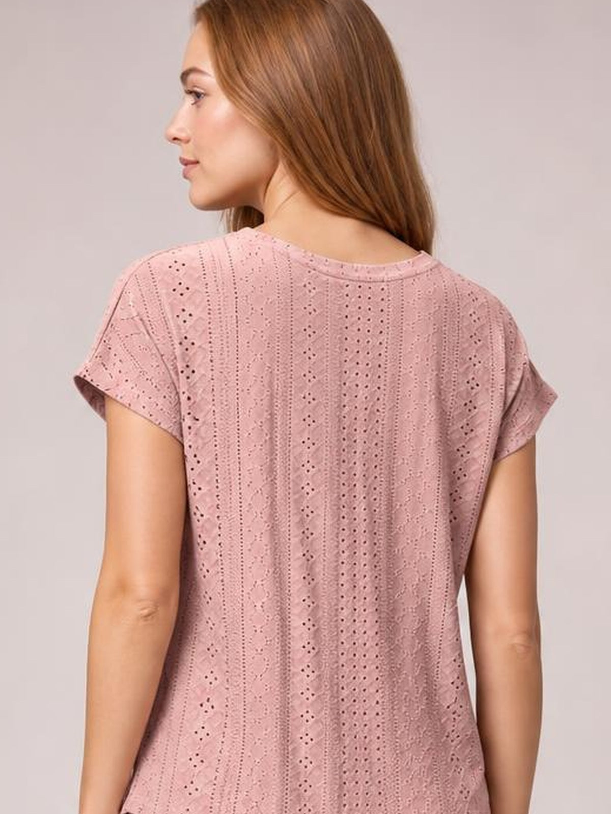 Back view of a model wearing a dusty pink eyelet short-sleeve top with rolled cuffs, vertical patterned construction, and a relaxed straight silhouette.