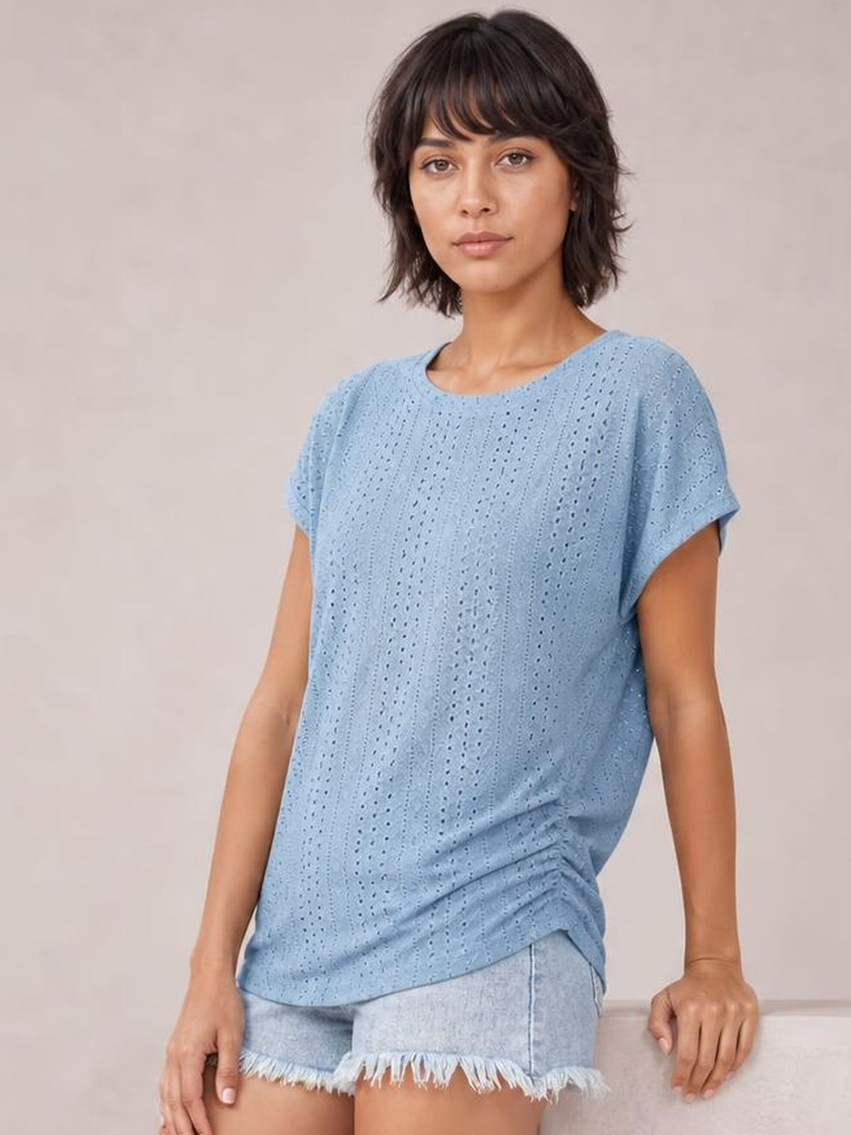 A model wears a light blue eyelet short-sleeve top with a relaxed silhouette, rounded neckline, softly draped hem, and delicate vertical openwork.