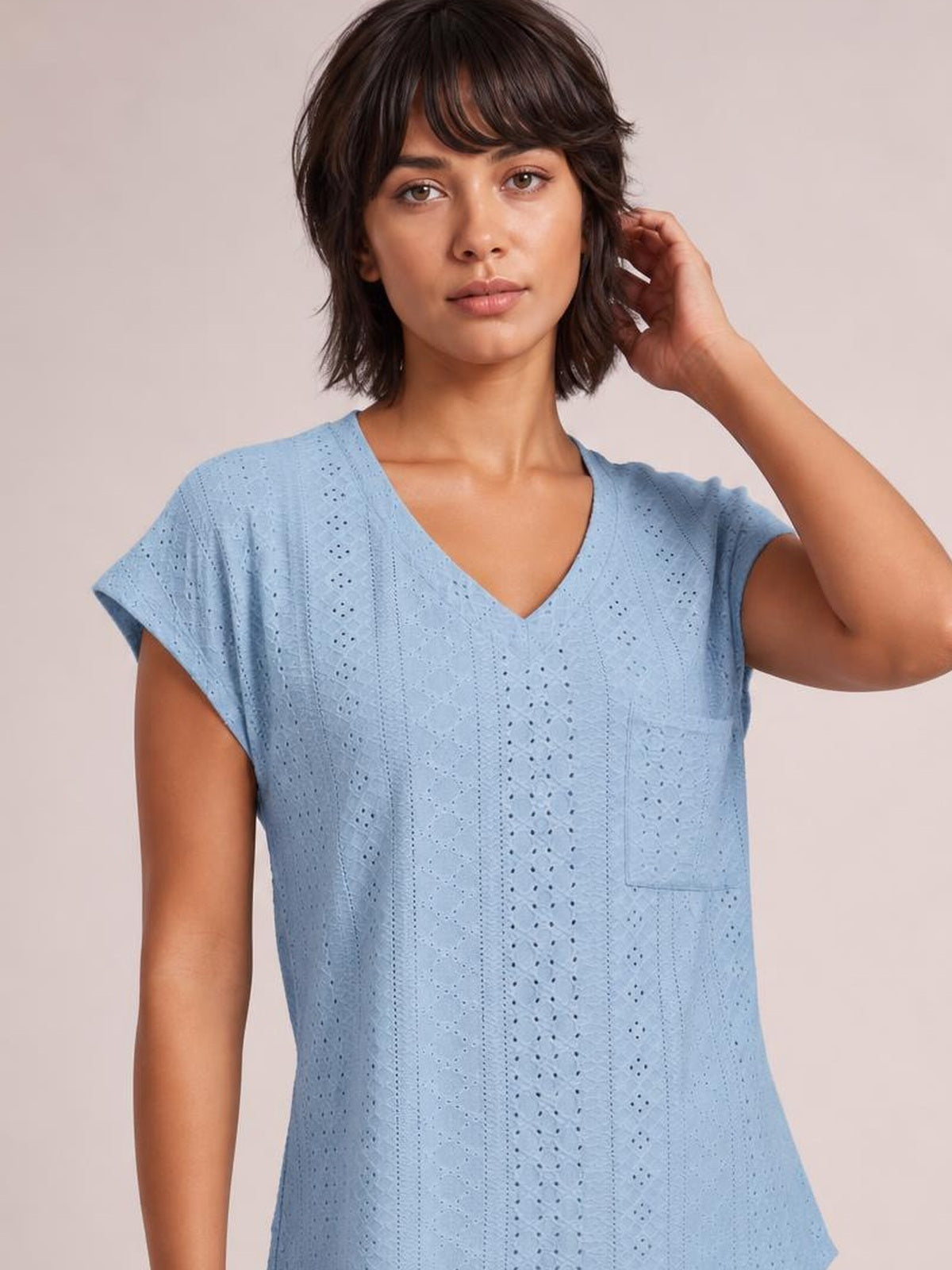 A model poses in a light blue eyelet V-neck short sleeve top with a relaxed straight silhouette and visible patch pocket, touching her hair.