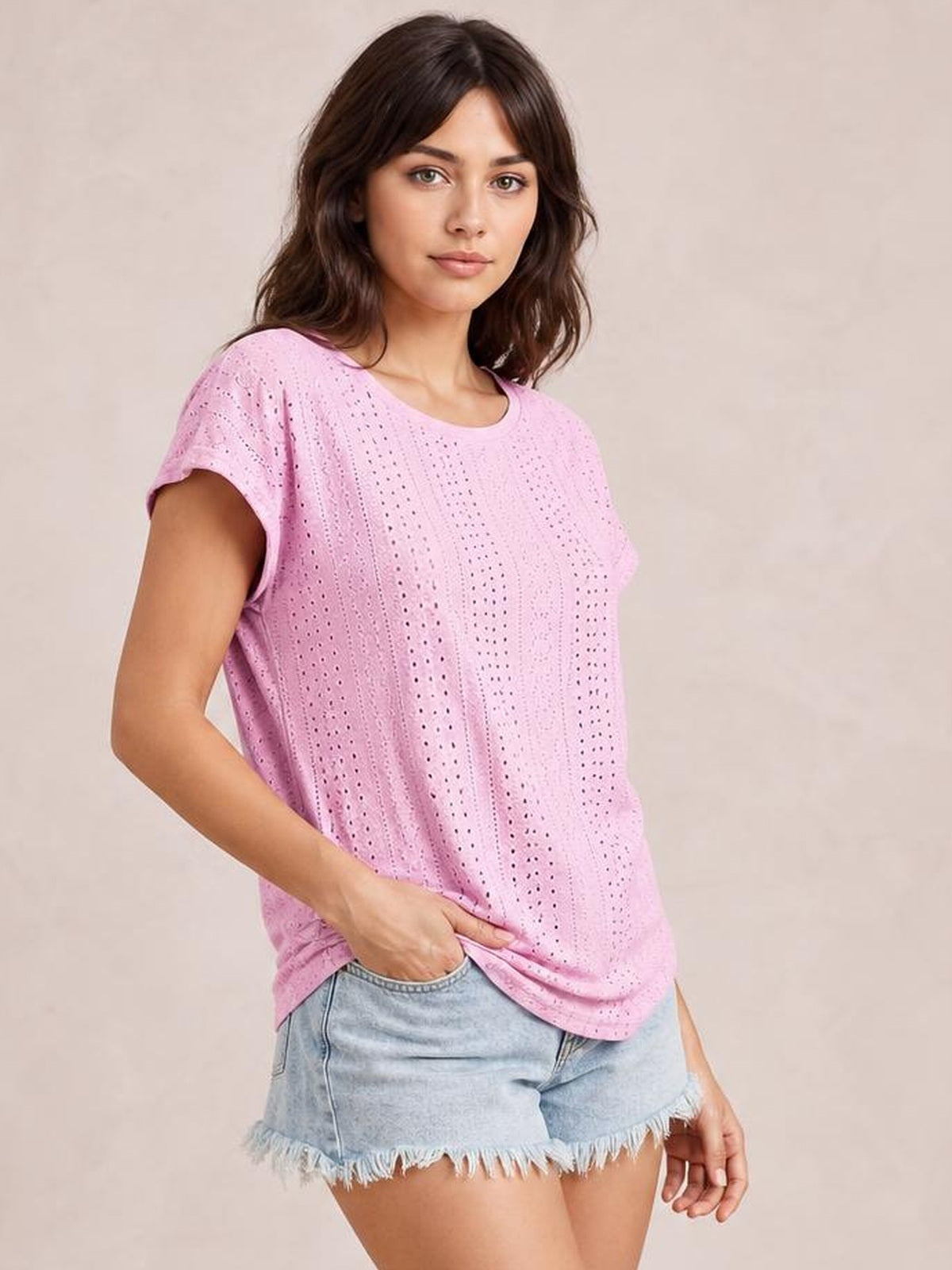 Model wearing a pink eyelet short-sleeve top with a relaxed silhouette and gently curved neckline, posing with one hand near the hem.