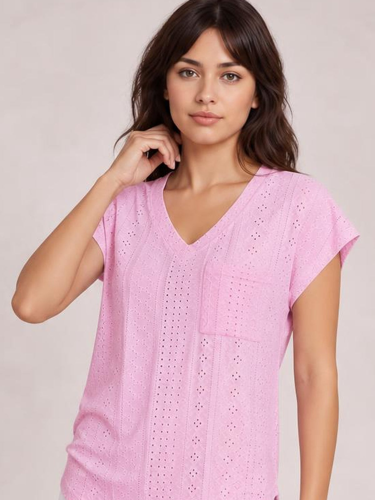 A model poses with one hand near her hair, wearing a pink eyelet V-neck short-sleeve top with a relaxed silhouette and visible chest pocket.