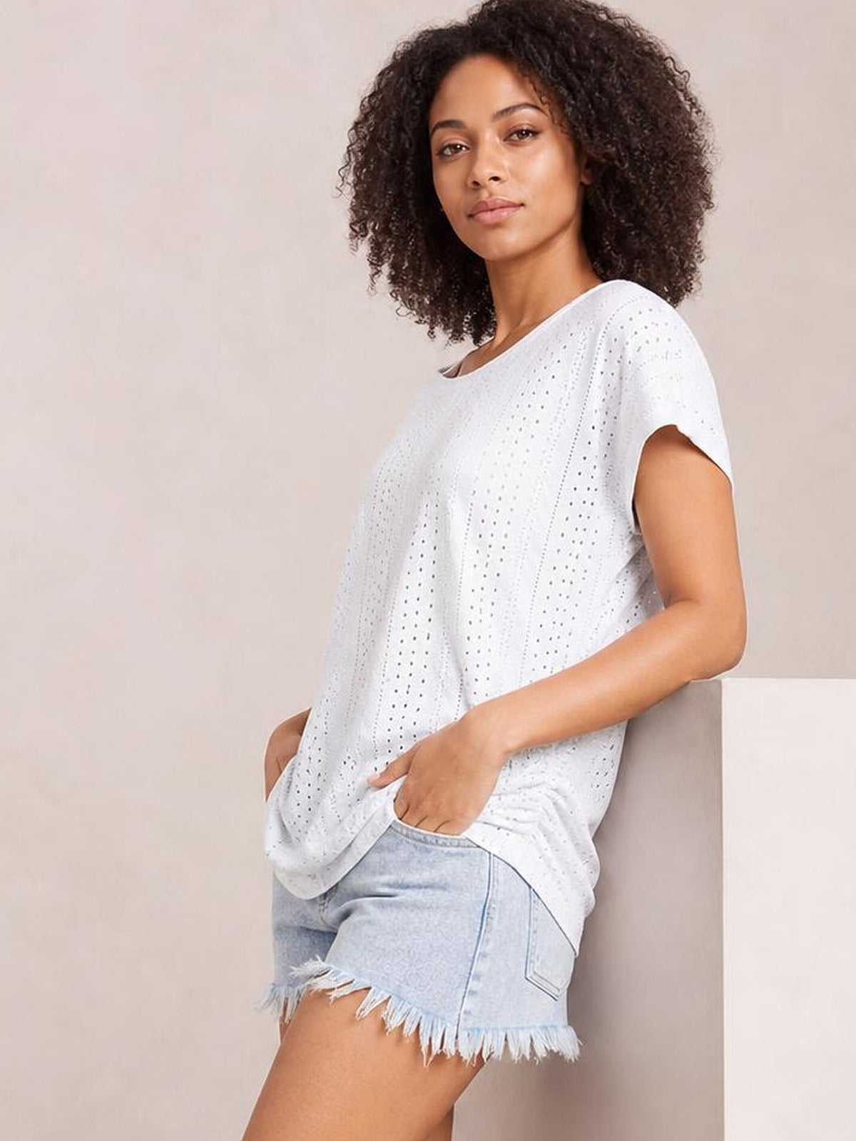 Model wears a white eyelet V-neck short sleeve top with a relaxed silhouette, vertical panel detailing, and practical side pockets, posing with hands tucked inside.