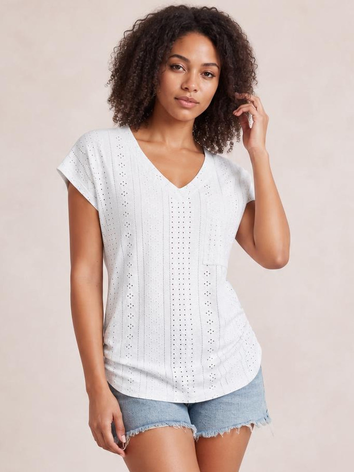 Model wearing a white eyelet V-neck short sleeve top with a relaxed silhouette, curved hem, and visible side pocket, posing with one hand in her hair.