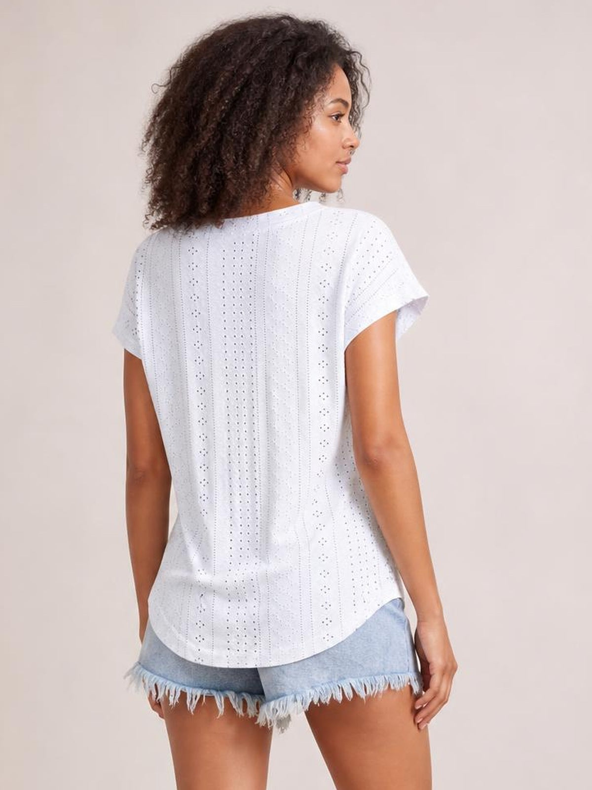 A model looks over her shoulder wearing a white eyelet short-sleeve top with a relaxed silhouette, vertical cutwork panels, and a curved hem.