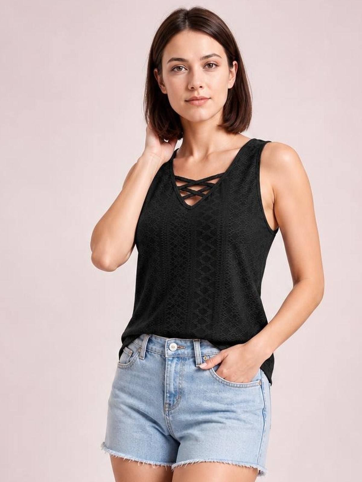 Model wears a black eyelet wide strap tank with a relaxed silhouette and crisscross neckline, posing with one hand in her denim shorts pocket.